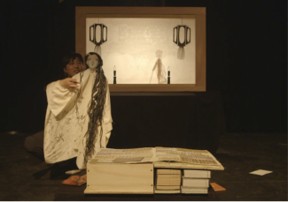 Shadow puppet screen in front of which is a puppeteer manipulating a Bunraku style puppet with a long white dress and hair