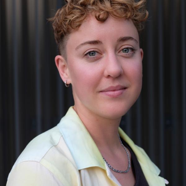 white person with short curly hair and a light colored collar shirt looking at the camera over their shoulder